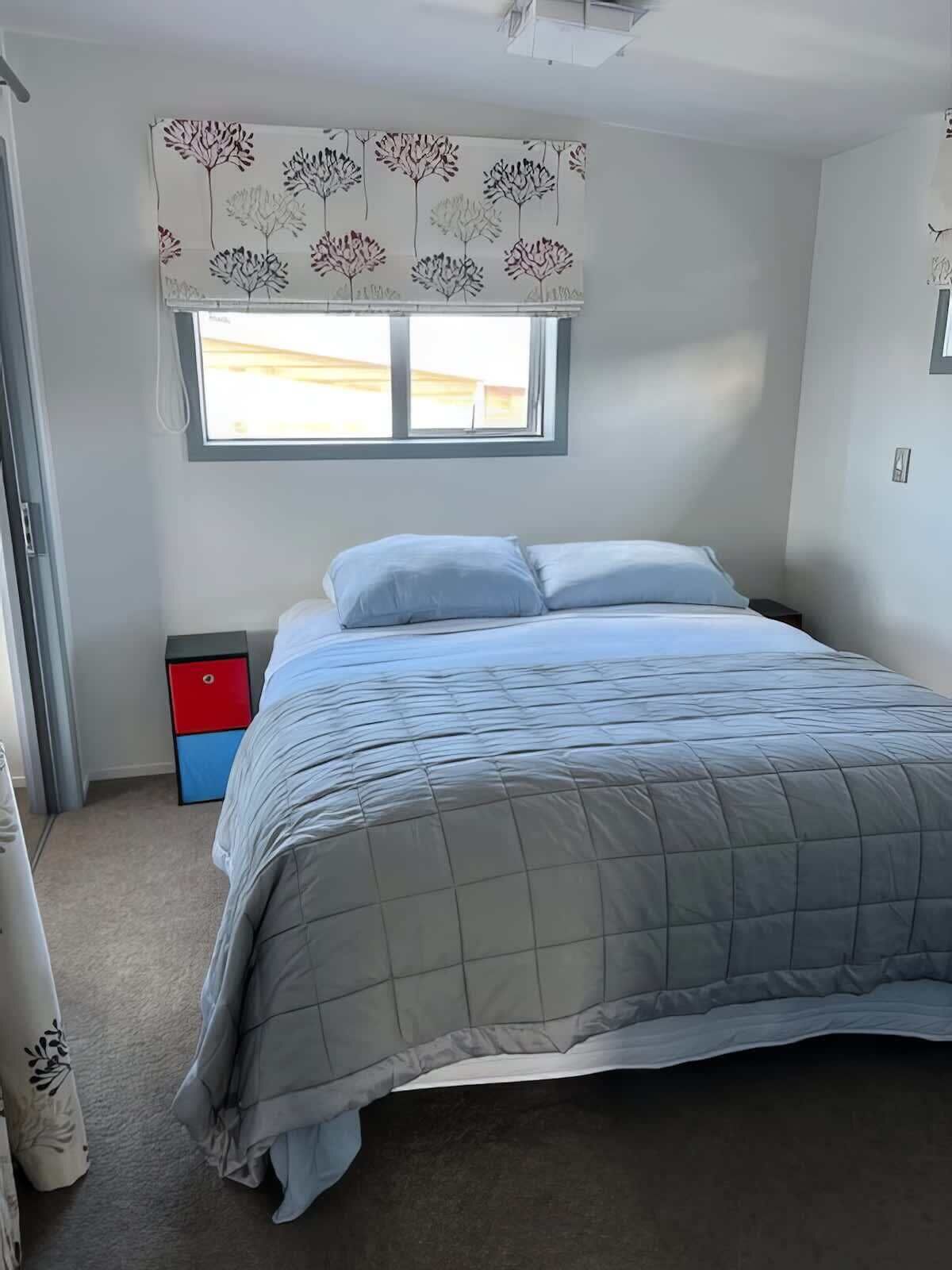 A bedroom with a bed, a window with a view, a nightstand, and a red and blue cabinet.