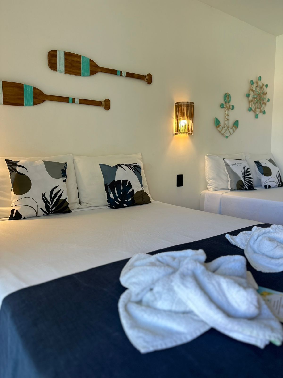 Une chambre d’hôtel avec deux lits, chacun recouvert d’une couvre-lit bleu foncé et agrémenté d’oreillers blancs à motifs de feuilles. Au-dessus des lits, sur le mur, on peut voir deux pagaies en bois, une lampe fixée au mur et une décoration murale.