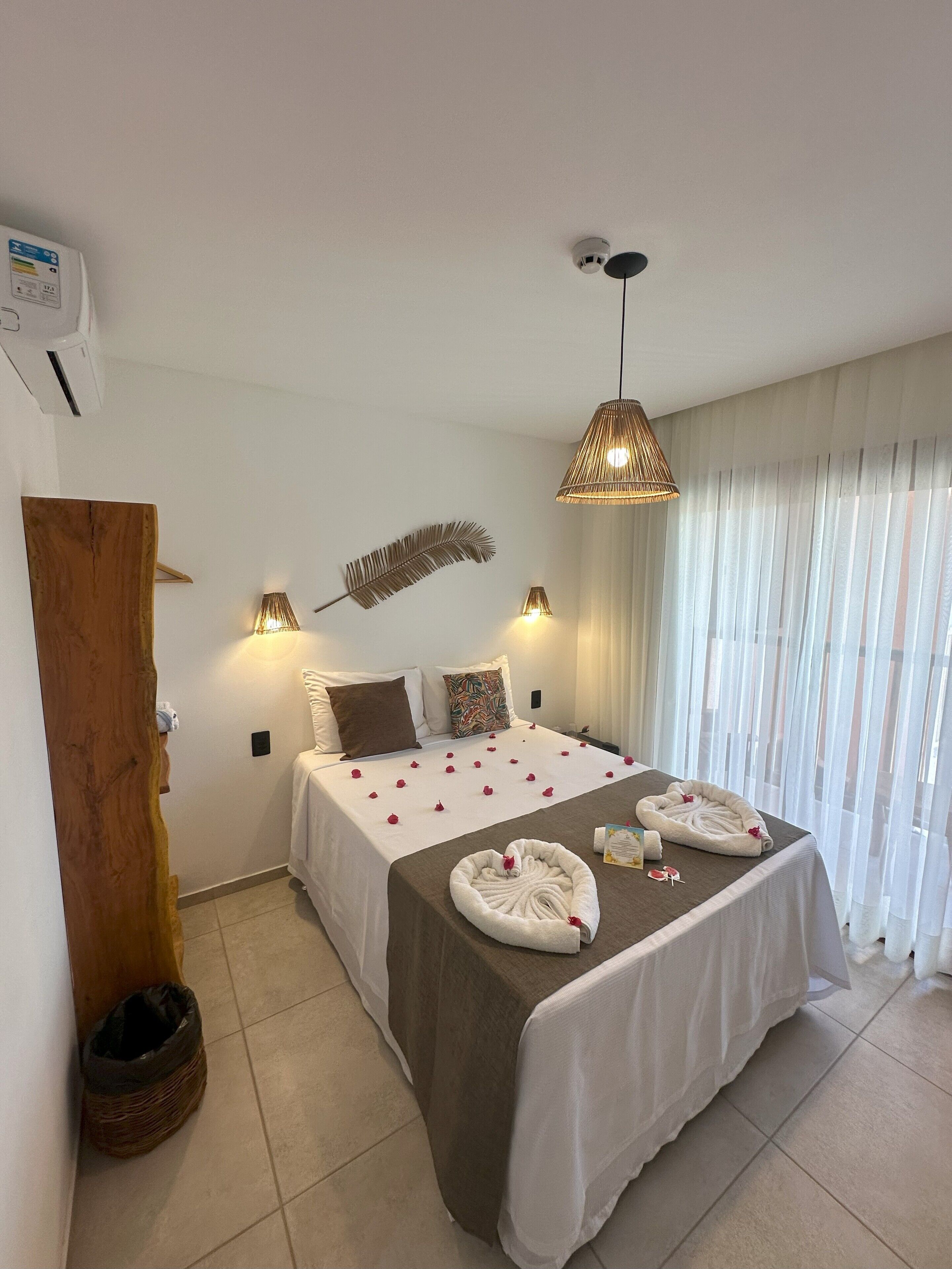A hotel room with a bed, two bedside lamps, a decorative feather wall hanging, a wooden trunk, and a window with sheer curtains.