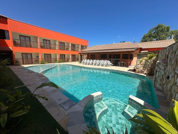 A swimming pool with a stone wall and a red building in the background. - Pousada Deu Praia Maragogi (Maragogi)