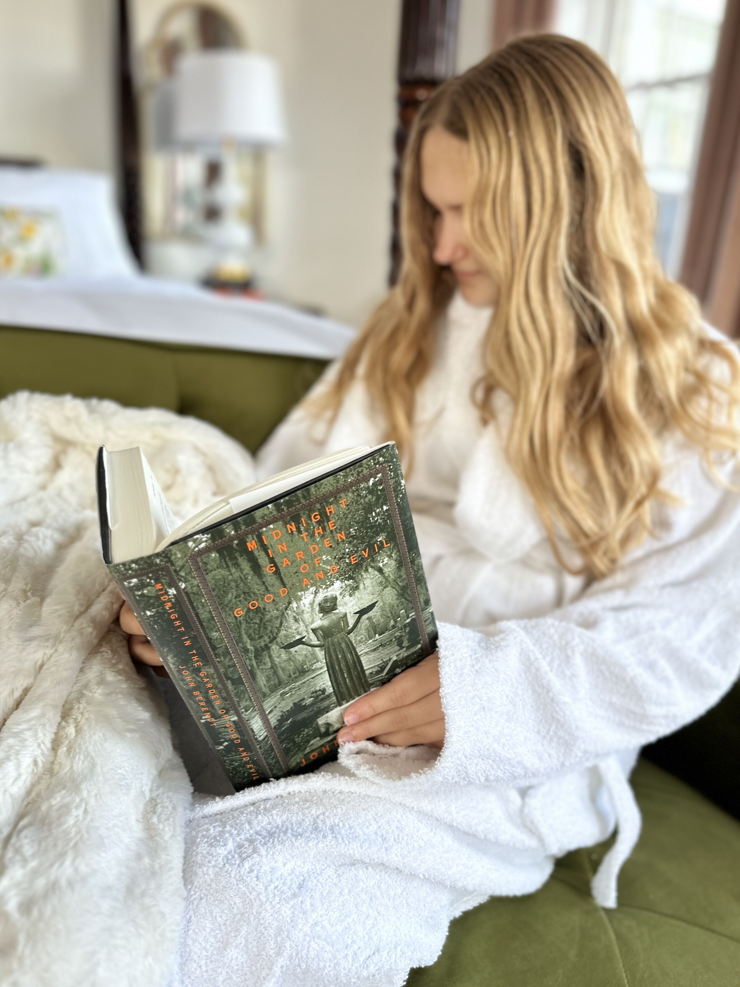 A person in a white bathrobe sitting on a green couch, reading a book titled "Midnight in the Garden of Good and Evil".
