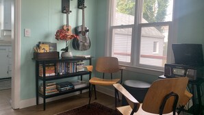 Ein Zimmer mit Bücherregal, zwei Gitarren, einem Sessel, einem Radio und einem Fenster mit Blick auf ein Haus.