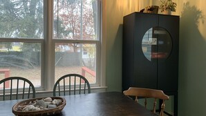 Ein Essbereich mit Tisch, Stühlen, einem Schrank und einem Fenster mit Blick auf Bäume und einen Zaun.
