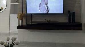 Een moderne woonkamer met een flatscreen televisie aan een houten wand, een decoratieve vaas met gedroogde bloemen en een klein plankje met een ingelijste foto en een vaas.