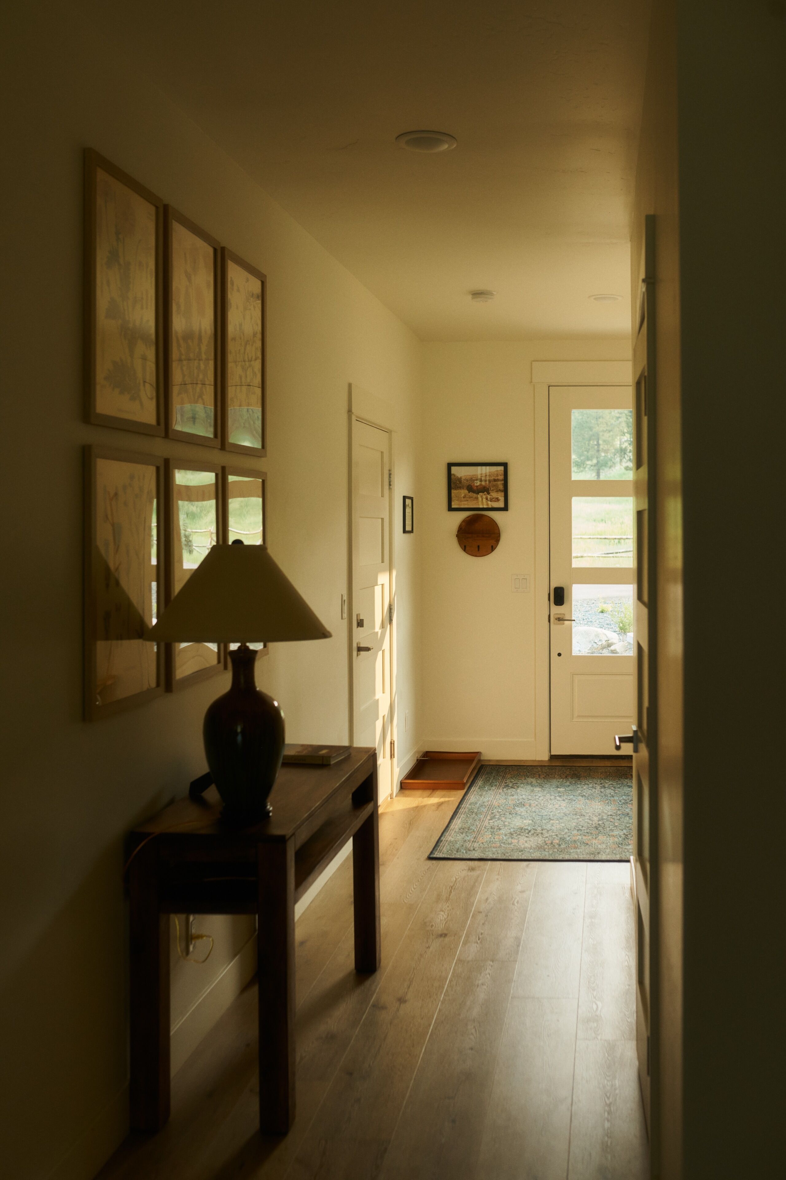 A hallway with a wooden floor, a table with a lamp, framed pictures on the wall, and a door with a window.