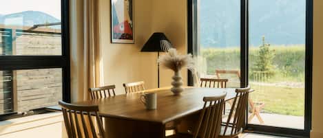 A dining area with a wooden table, chairs, and a vase. There is a window with a view of mountains and a door leading outside.