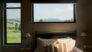 Een slaapkamer met een groot raam, een bed met veel kussens, twee nachtkastjes met lampen en uitzicht op een groen landschap.