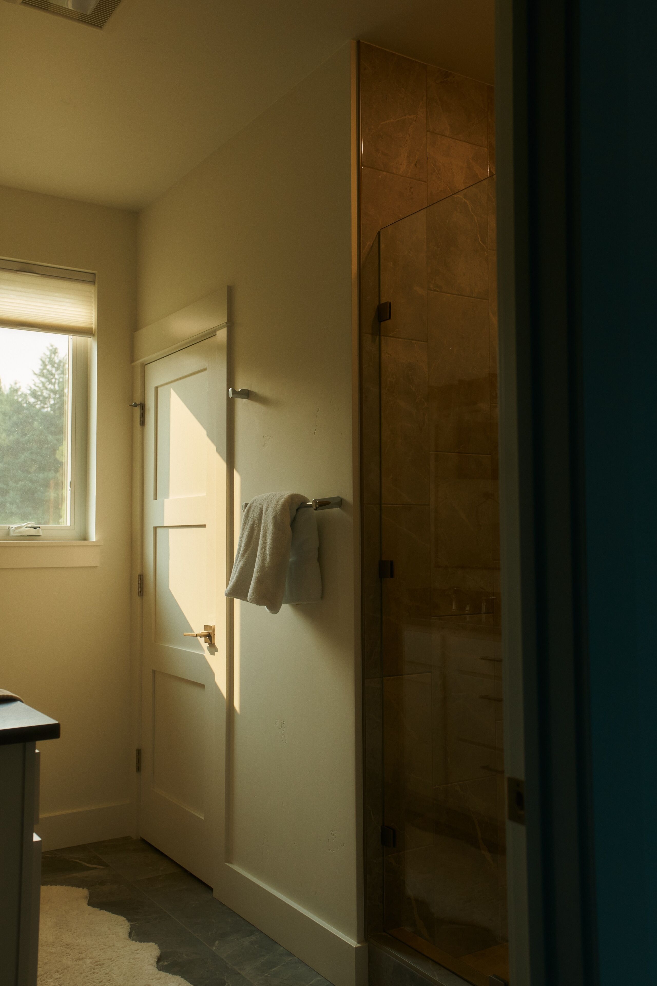 A bathroom with a glass shower enclosure, a towel hanging on the wall, and a window providing natural light.