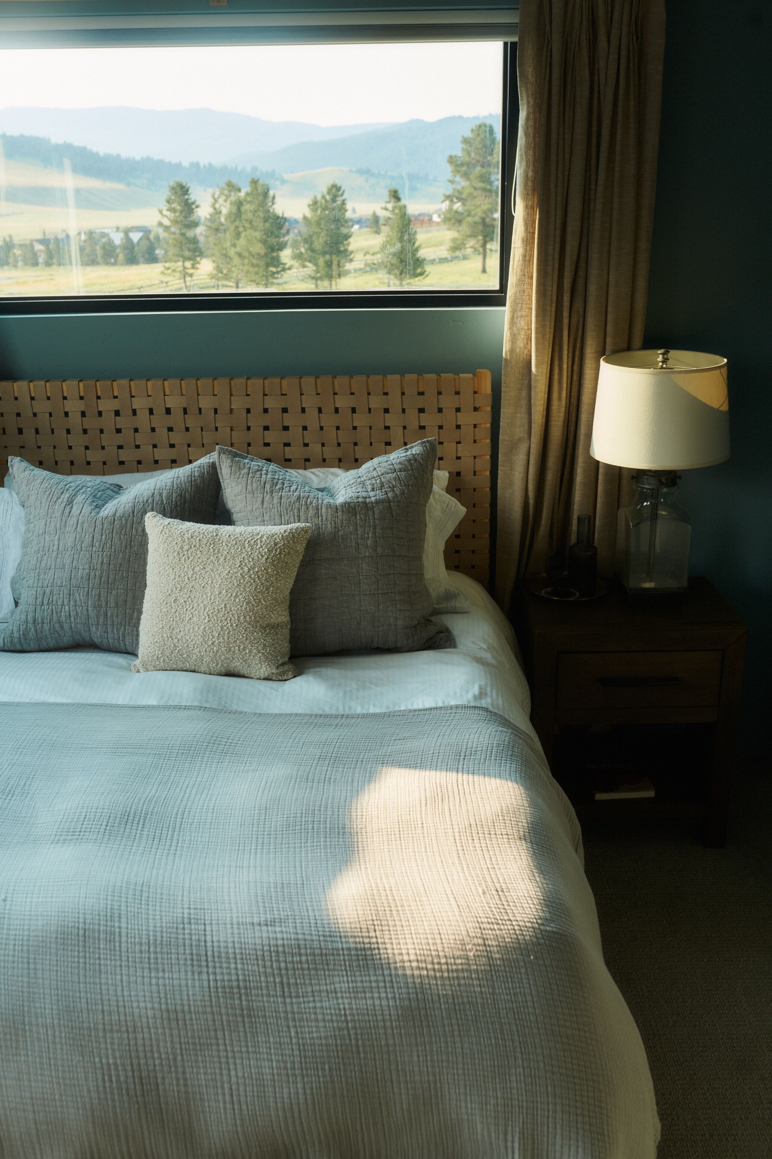 A neatly made bed with a view of a landscape through a window, a bedside lamp, and a wooden headboard.