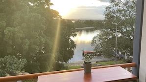 A balcony with a wooden table and two white stools, overlooking a lake with trees and a dock. - Water views in town centre. Secure parking with lift. (Forster)