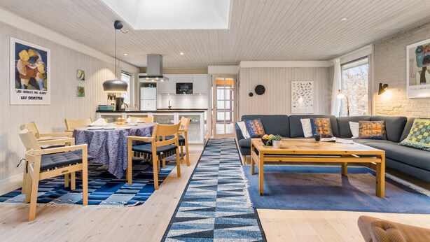 A modern living room with a sofa, coffee table, and dining area. There is a kitchen in the background, a skylight above the dining area, and artwork on the walls.