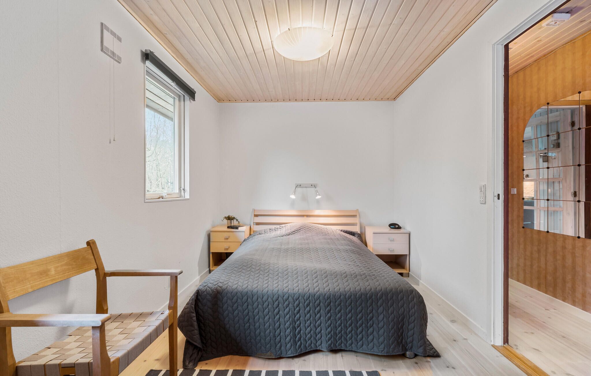 A bedroom with a bed, bedside tables, a chair, a window, and a wooden ceiling.