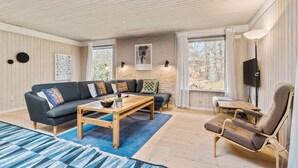 A cozy living room with a sofa, coffee table, and a chair. There are two windows, a TV, and artwork on the walls.