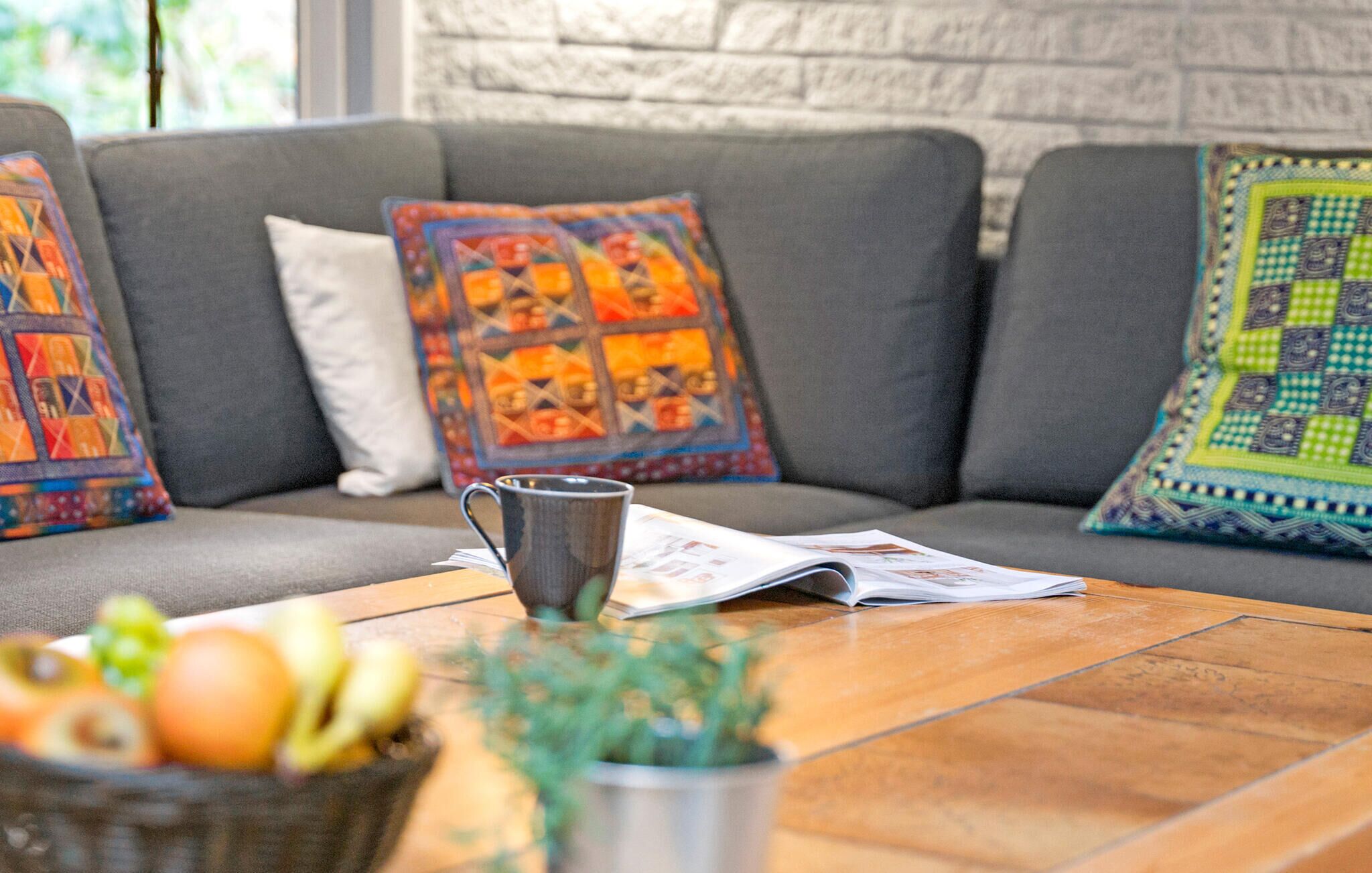Un salon accueillant avec un canapé gris agrémenté de coussins colorés, une tasse en métal, un journal et un saladier de fruits posés sur une table basse en bois.