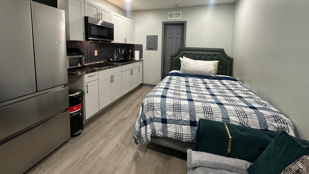 A compact room with a kitchenette, a bed with a checkered duvet, a ceiling fan, and a door.