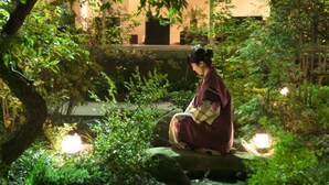 Una persona sentada sobre una roca en un jardín con plantas y un edificio al fondo.