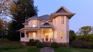 Une maison à deux étages dotée d’un porche qui fait le tour de la maison, d’une entrée centrale avec des marches et d’une grande fenêtre au deuxième étage.