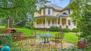 Une maison à deux étages dotée d’un porche, d’une cheminée en pierre et d’un jardin agrémenté de chaises et de tables en métal.