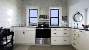Une cuisine moderne avec des meubles blancs, une cuisinière en acier inoxydable et un four à micro-ondes. Deux fenêtres laissent entrer la lumière naturelle, et une horloge est fixée au mur.