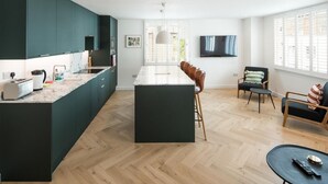 A modern kitchen with dark green cabinets, a marble countertop, and a central island. There is a dining area with a sofa and chairs, a TV mounted on the wall, and a large window with blinds. - 2Bed 2Bath Stunning Shoreditch Apartment (London)