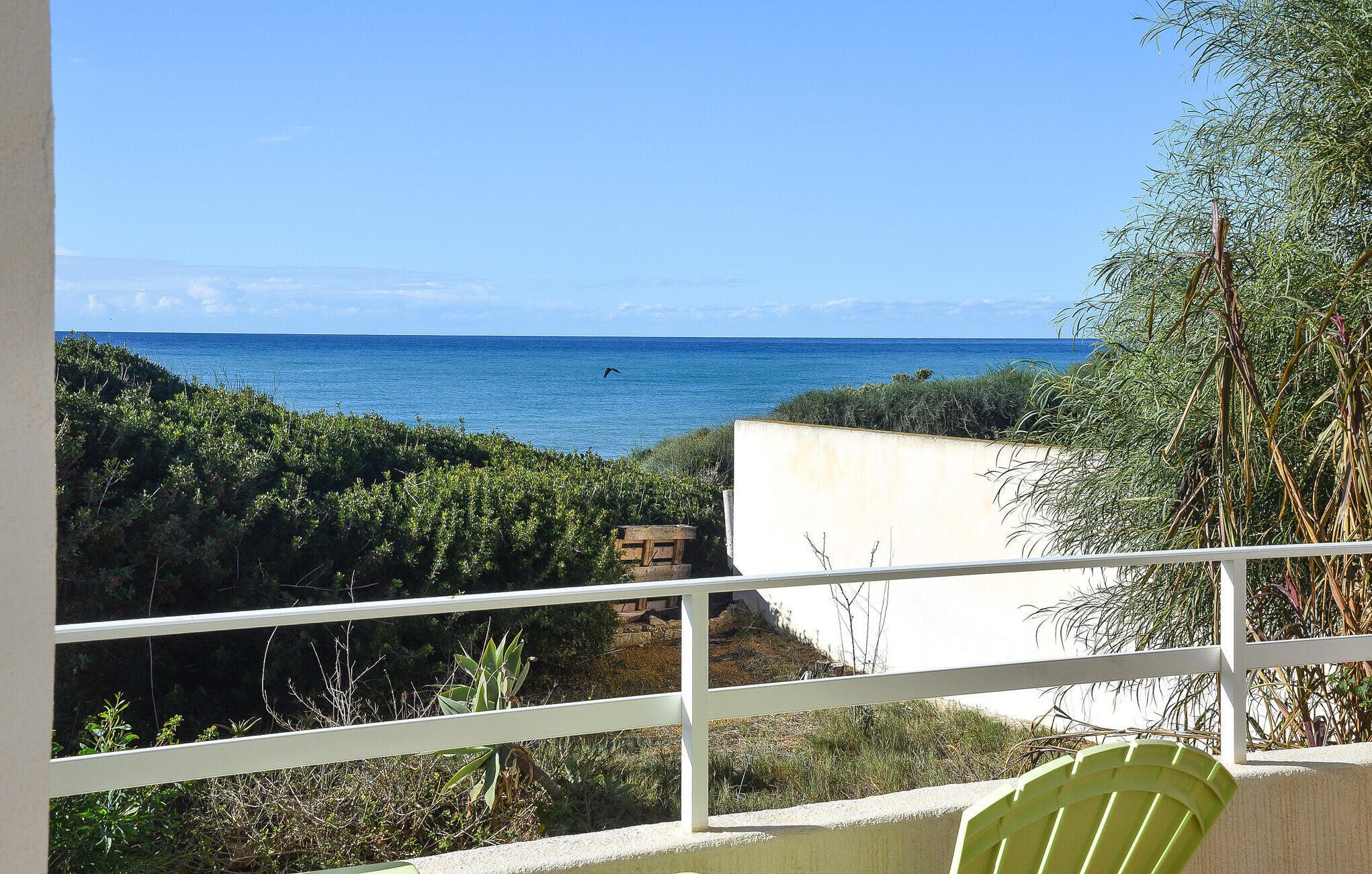 A balcony with a view of the sea, surrounded by greenery and a white building.