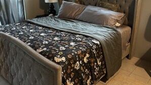A bedroom with a tufted headboard, a floral bedspread, a nightstand with a lamp, and a window with a geometric pattern. - Bay Bliss Retreat home in Ironshore, Montego Bay, less than 10 mins to airport. (Montego Bay)