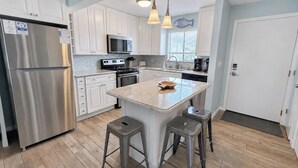 A modern kitchen with white cabinets, stainless steel appliances, a granite countertop island, and two bar stools. - Snowbird Retreat - Private Beach, Heated Pools, Pickleball & Golf! (Destin)