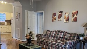 A living room with a plaid sofa, wooden flooring, a ceiling fan, and a door leading to a kitchen. - Lake House on Weiss Lake! (Leesburg)