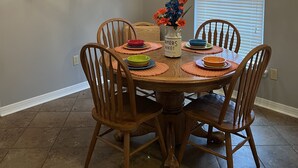A dining area with a round wooden table, six chairs, a chandelier, a floral window valance, a colorful abstract painting, and a vase with flowers. - Lake House on Weiss Lake! (Leesburg)