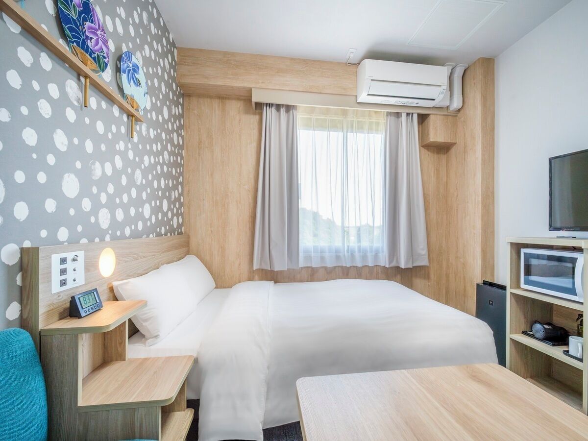 A hotel room with a bed, bedside table, TV, and an air conditioning unit.
