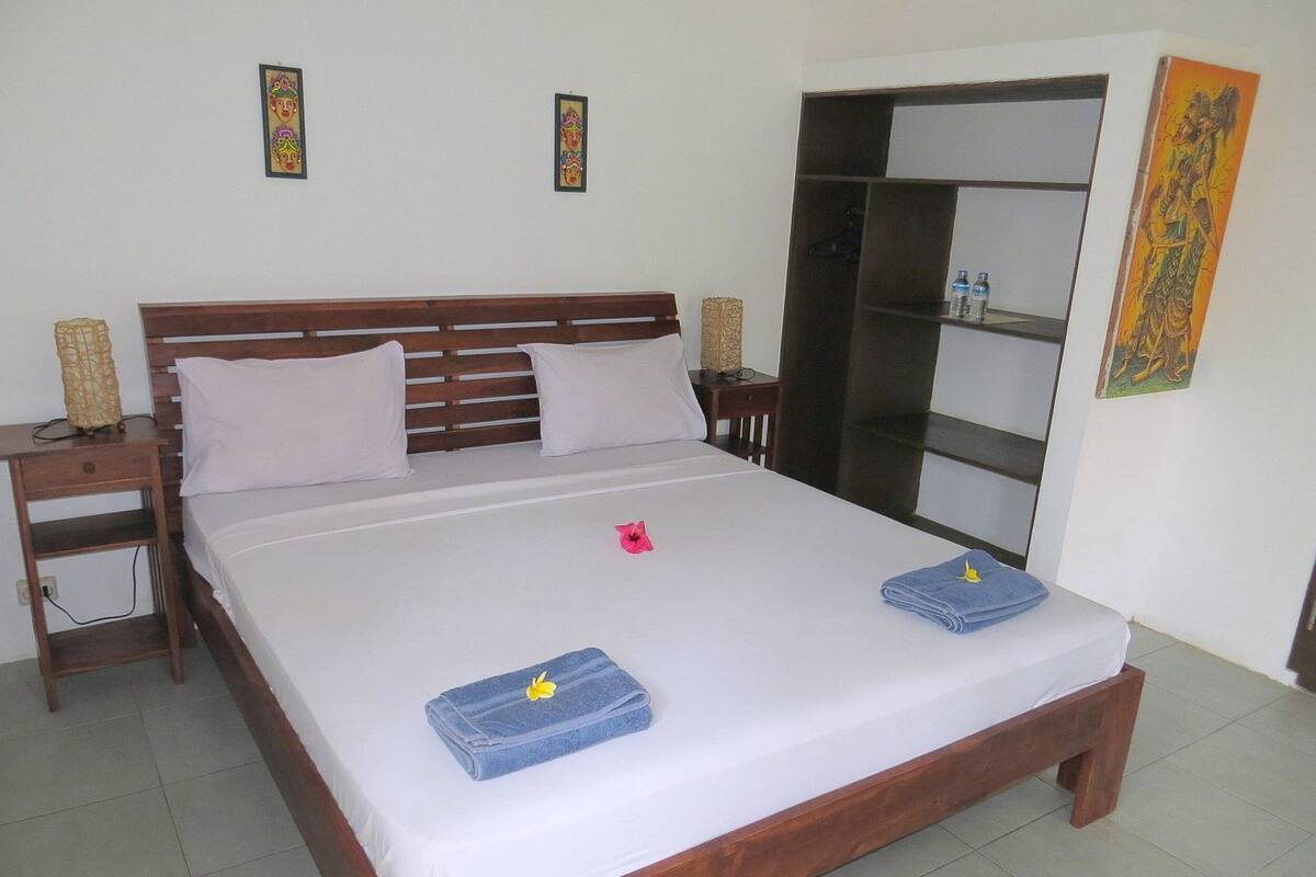 A neatly made bed with white linens, two pillows, and two folded towels with yellow flowers. There is a wooden nightstand with a lamp, a small table with a bottle, and a wardrobe with a mirror.