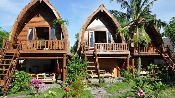 Two wooden cottages with balconies, surrounded by tropical vegetation and palm trees.