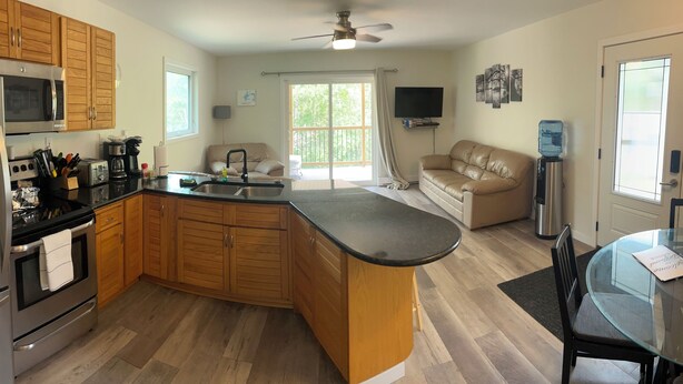 A modern kitchen with wooden cabinets, a stainless steel refrigerator, a microwave, and a coffee maker. There is a dining area with a glass table and chairs, and a sofa in the living room. - 4 Season Cabin great for Summer or Winter activities! 
Excellent Green Space (Marean Lake)