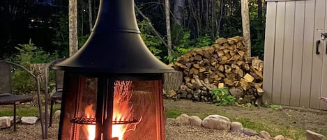 A wood-burning stove with a chimney, surrounded by a gravel area, a stack of firewood, and string lights in a wooded setting.