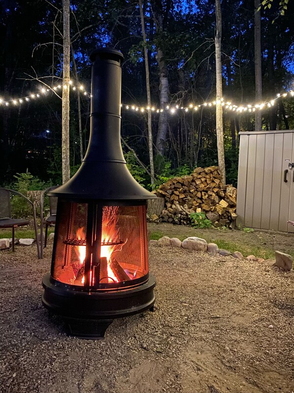 A wood-burning stove with a chimney, surrounded by a gravel area, a stack of firewood, and string lights in a wooded setting.