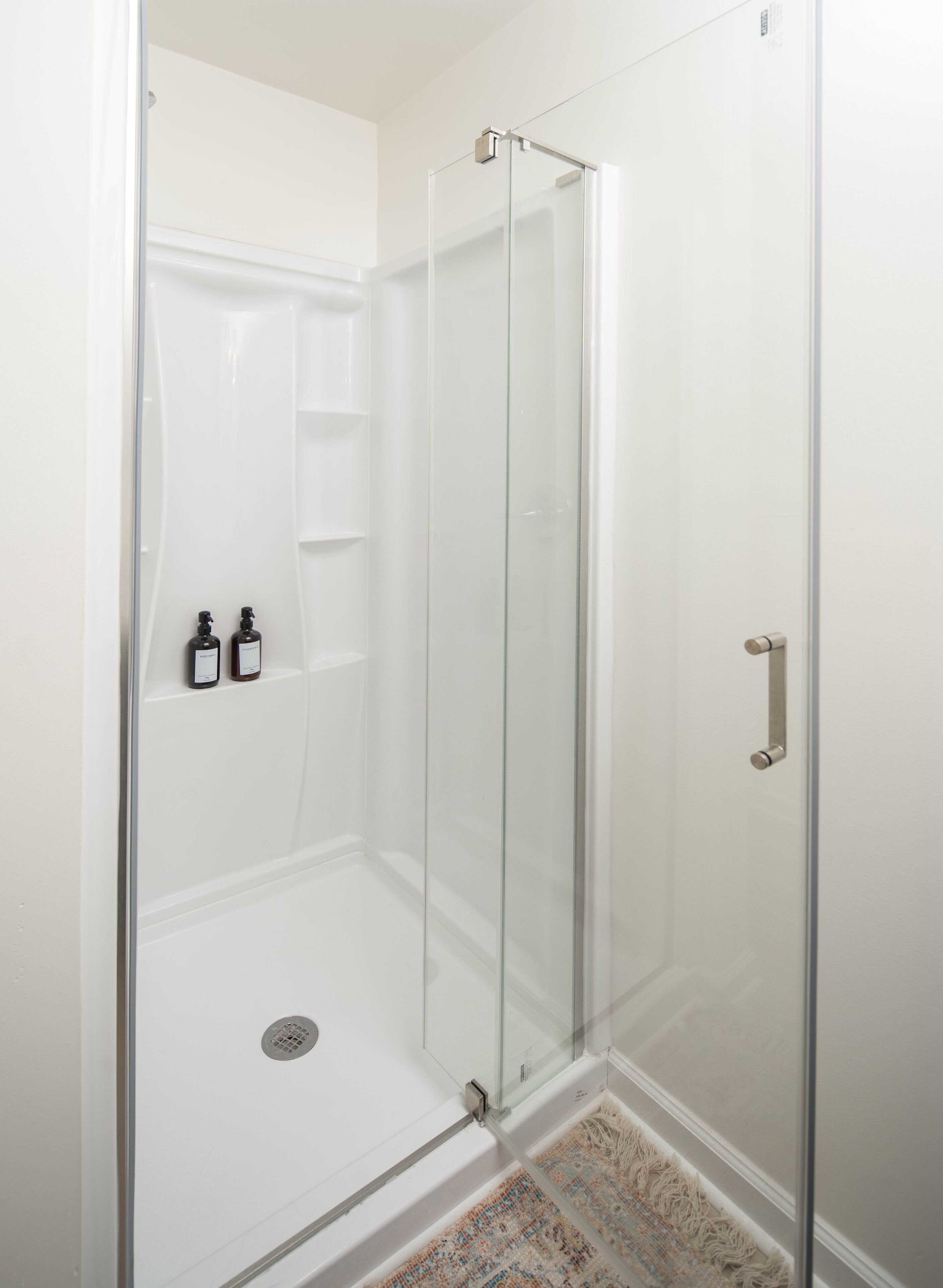 A glass-enclosed shower with a white interior, a metal handle, and two bottles of shampoo on a shelf.