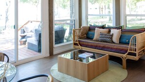 Une pièce spacieuse avec une grande fenêtre, un ventilateur de plafond, un canapé avec des coussins, une table basse en verre et un fauteuil en osier.