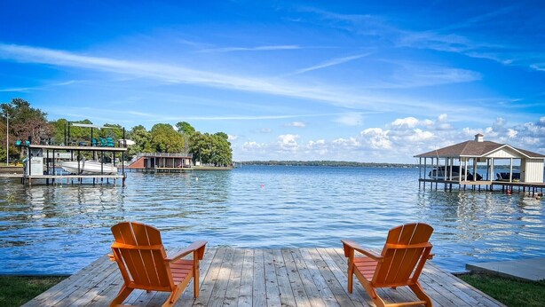 Une terrasse en bois avec deux fauteuils Adirondack en bois, donnant sur un lac paisible, avec des quais et des bâtiments en arrière-plan.
