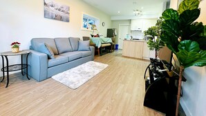 Una sala de estar moderna con un sofá gris, una mesita redonda y un tapete blanco. Se observa una zona de cocina con armarios blancos y una planta verde grande.