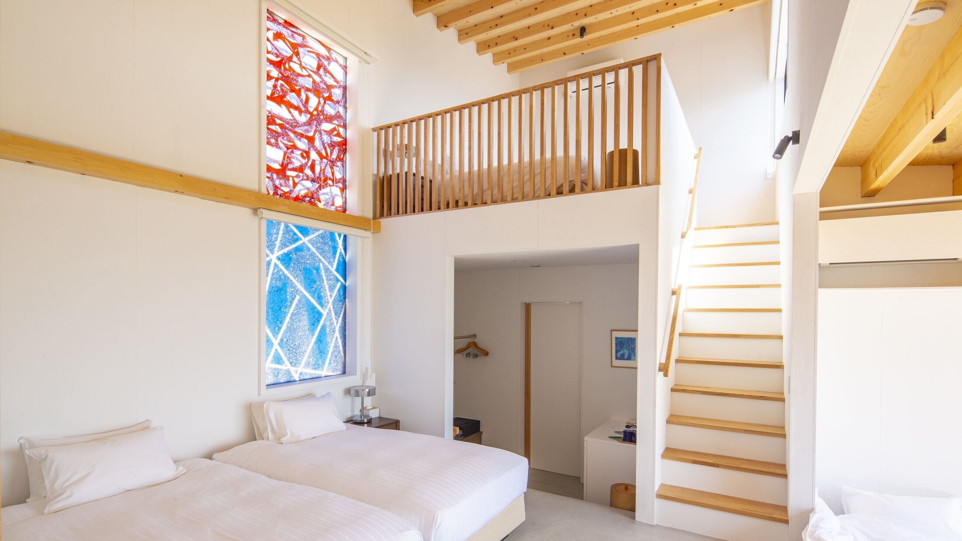 A modern bedroom with a bed, a window with a geometric pattern, a wooden staircase, and a door leading to another room.