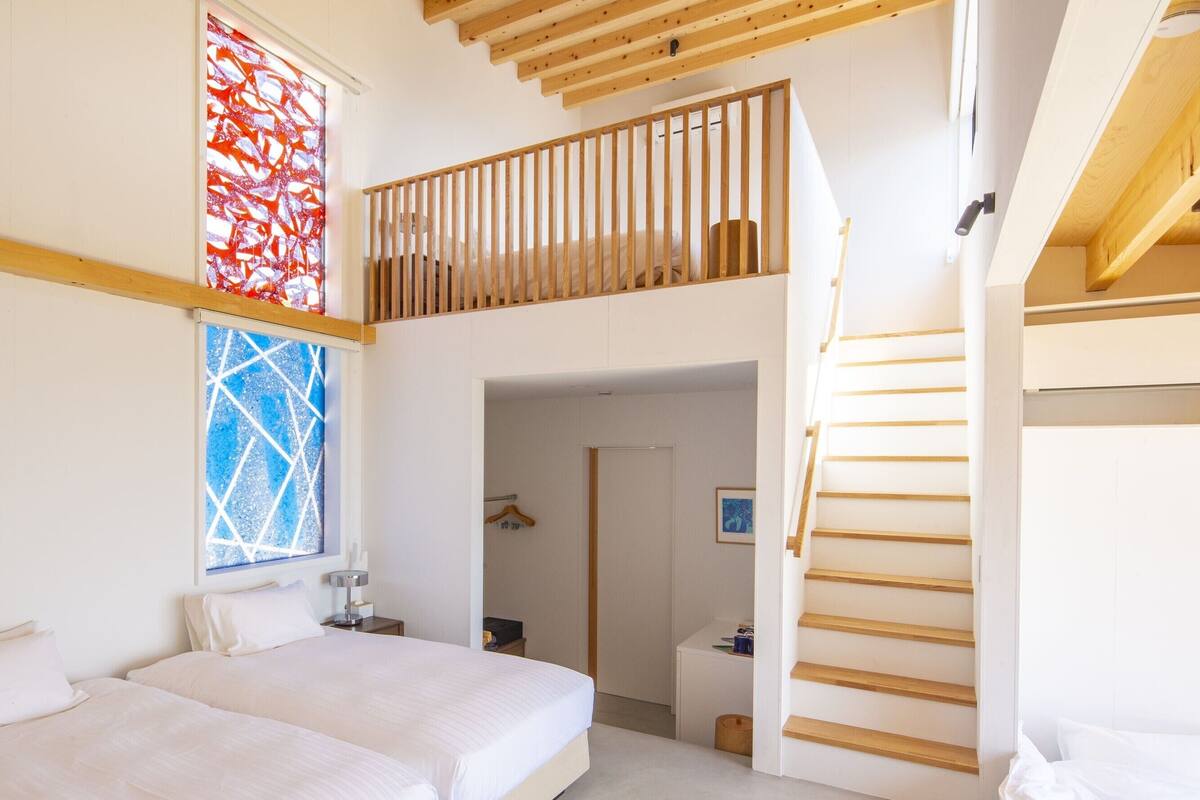 A modern bedroom with a bed, a window with a geometric pattern, a wooden staircase, and a door leading to another room.