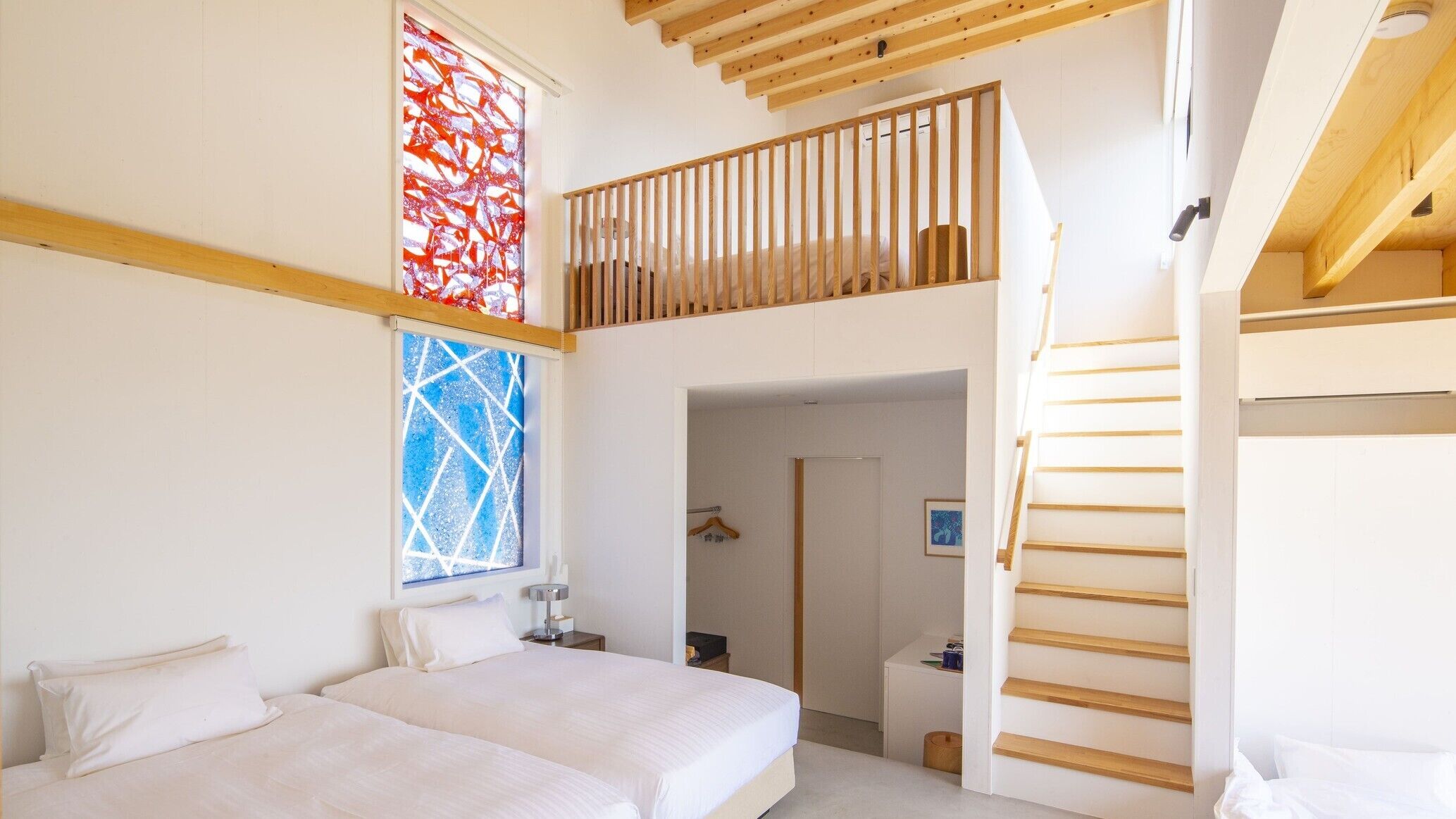 A bedroom with a bed, a window with a geometric pattern, a wooden staircase, and a door leading to another room.