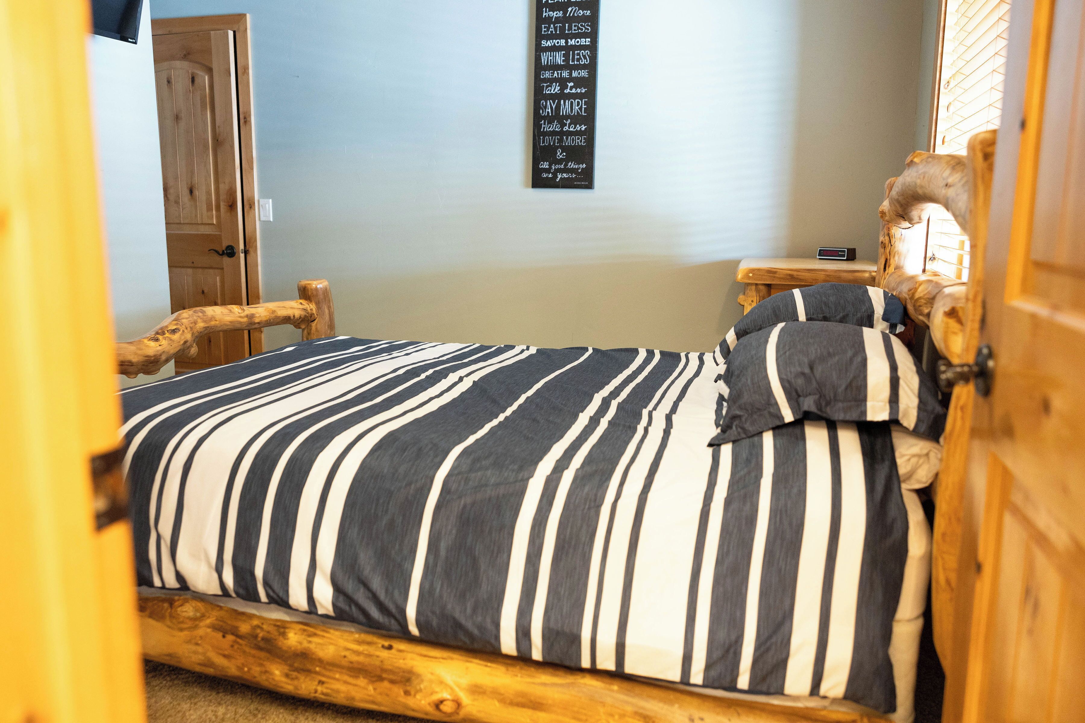 Un dormitorio con estructura de cama de madera, ropa de cama a rayas, una mesita de noche pequeña, un oso de peluche y una cita motivacional en la pared.