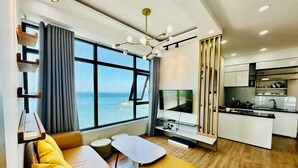Una sala moderna con un sofá, una mesita y un televisor. Se observa una zona de cocina con armarios y un lavamanos.