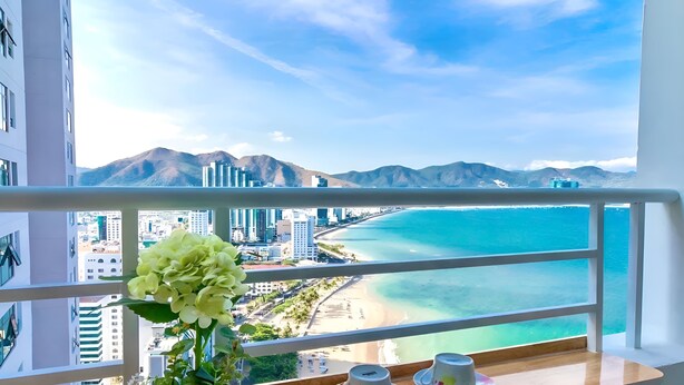 Un balcón con vistas a una playa y montañas, una planta en maceta y dos tazas sobre platillos.