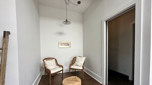 A narrow hallway with two wooden chairs, a round ottoman, a ceiling light, and a framed picture on the wall.