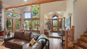 A spacious living area with a leather sofa, wooden staircase, and large windows offering views of the outdoors.