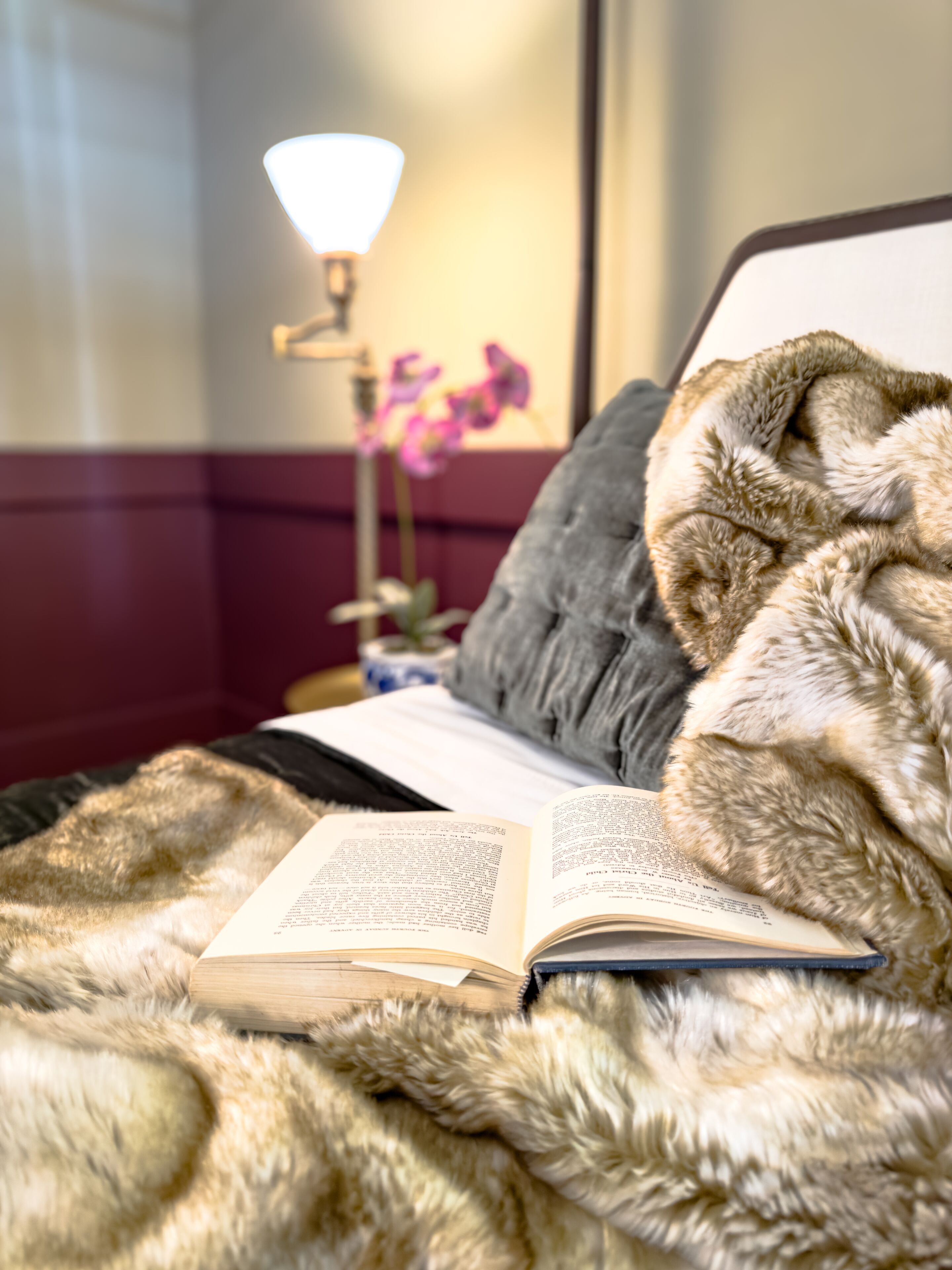A bed with a fur blanket, an open book, and a bedside lamp.