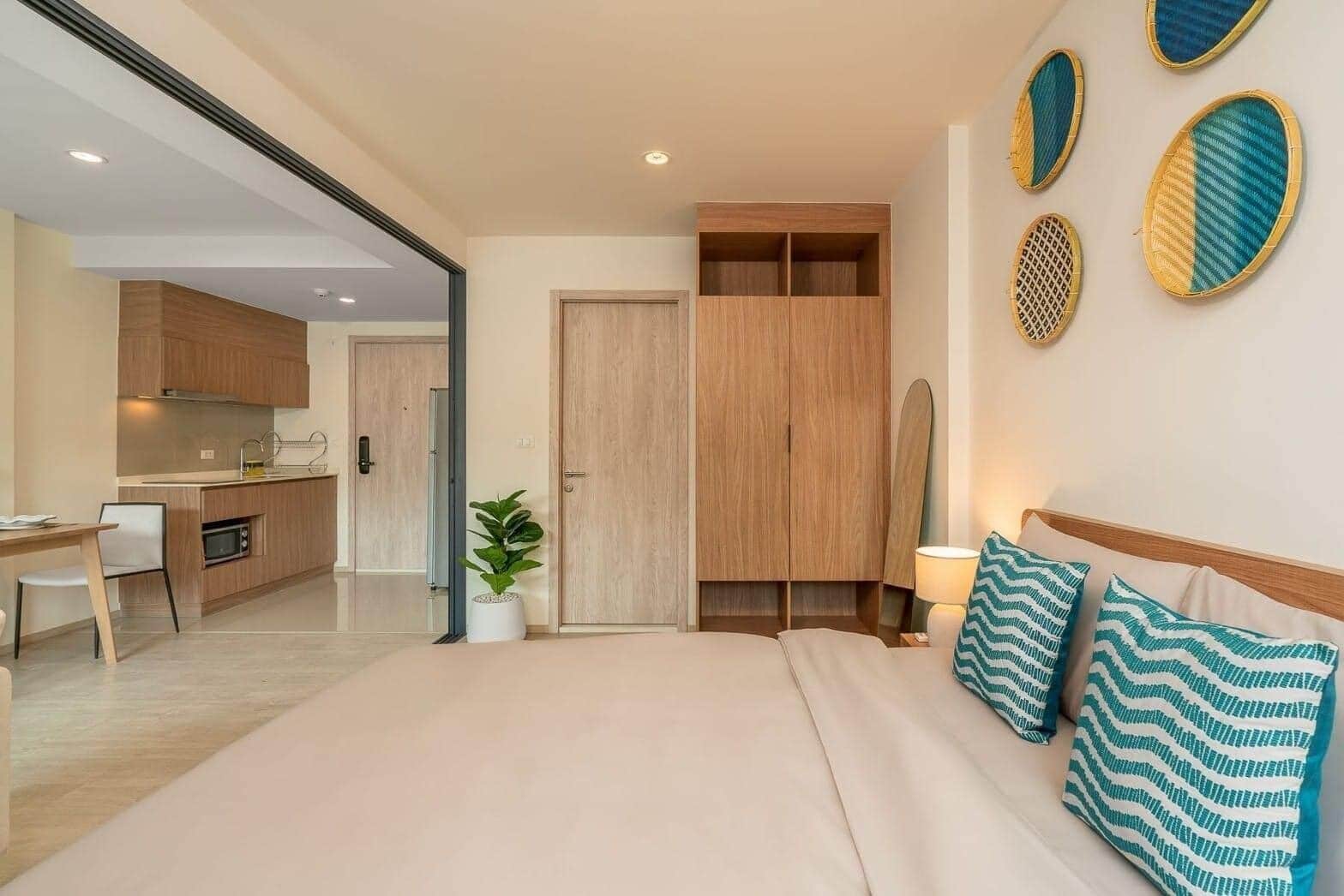 Une chambre moderne avec un grand lit, une table de chevet et une commode. On aperçoit un placard en bois et un petit coin cuisine à travers une porte ouverte.