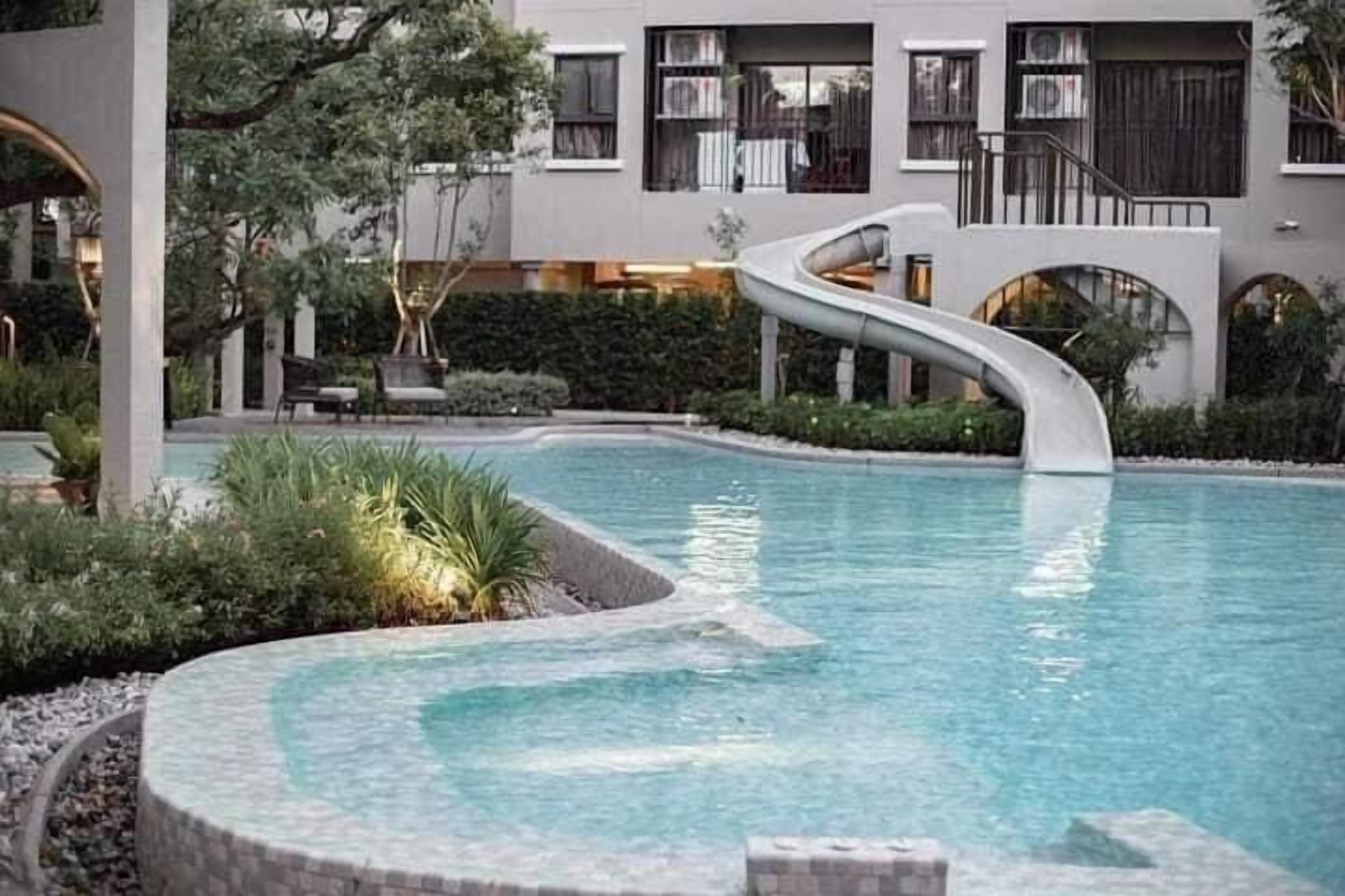 Een modern zwembad met een uniek, gebogen ontwerp, omringd door een aangelegde tuin en een gebouw met balkons.